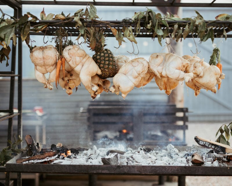 Whole chicken roasting over flames and hot coals with smoke rising.